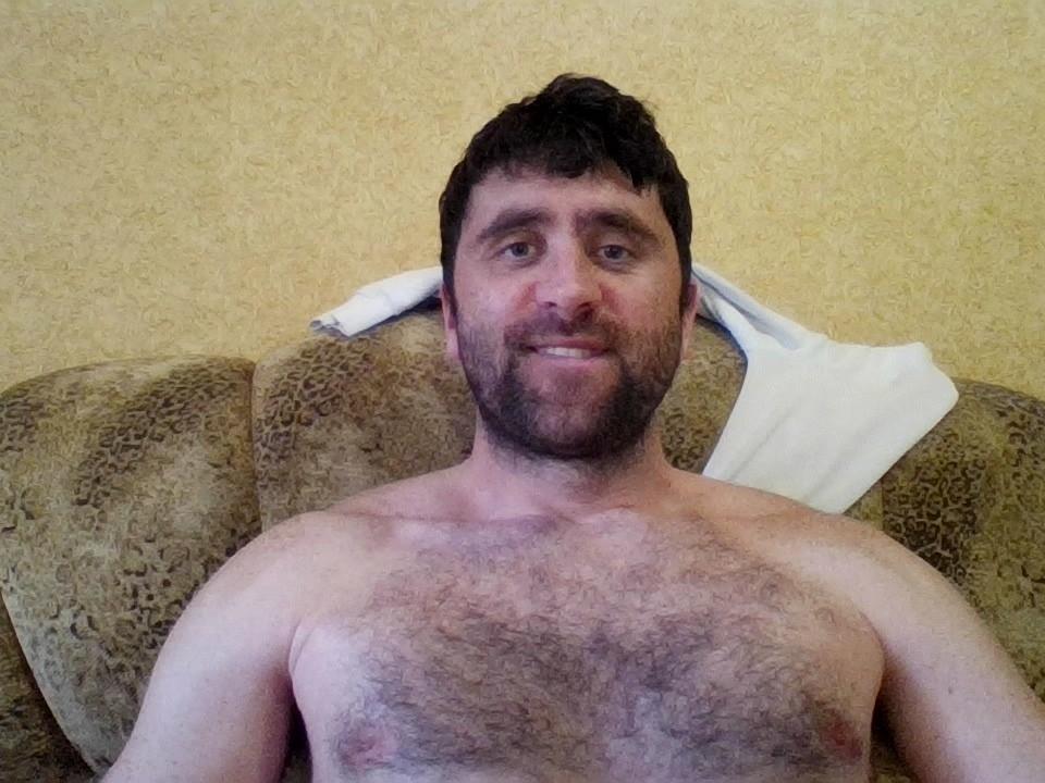 StasConnor, sem camisa, com o peito peludo, cabelo curto e escuro, e barba, sorrindo para a câmera, apoiado em um sofá estampado com uma toalha branca atrás da cabeça..