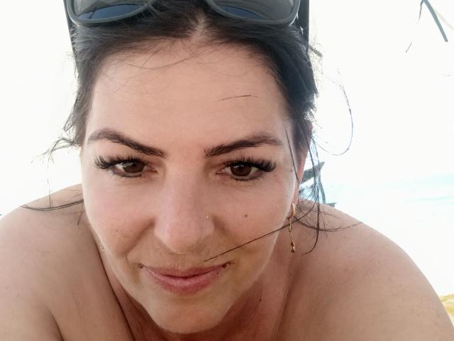 YulliaMilfX has dark brown eyes, long eyelashes, and a small nose piercing. She's topless with sunglasses on her head, a slight smile, and a thin hair strand on her cheek.
