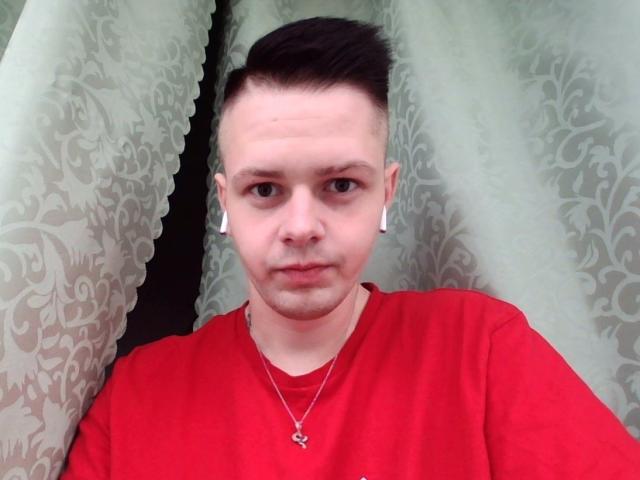 MikeXKing heeft kort donker haar, witte oordopjes, een rood shirt en een zilveren ketting met een klein kruisje. Hij kijkt recht in de camera met een neutrale uitdrukking..