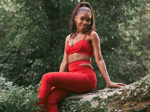 AlanaPatel, with dark skin and curly hair in a ponytail, sits on a tree branch in red sports bra and pants, smiling at the camera, showing her small ass.