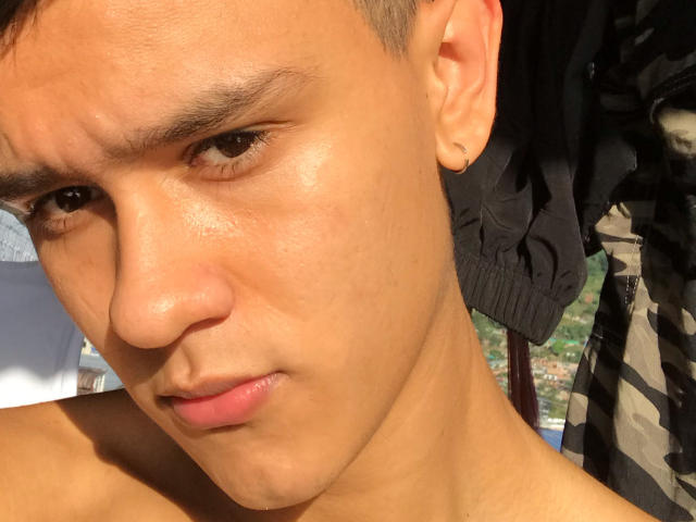 LoganD a un visage bronzé et brillant, avec des cheveux courts et bruns, de petites oreilles, et une seule piercing à l'oreille, il regarde vers le bas avec une expression légèrement confuse, et il est torse nu..