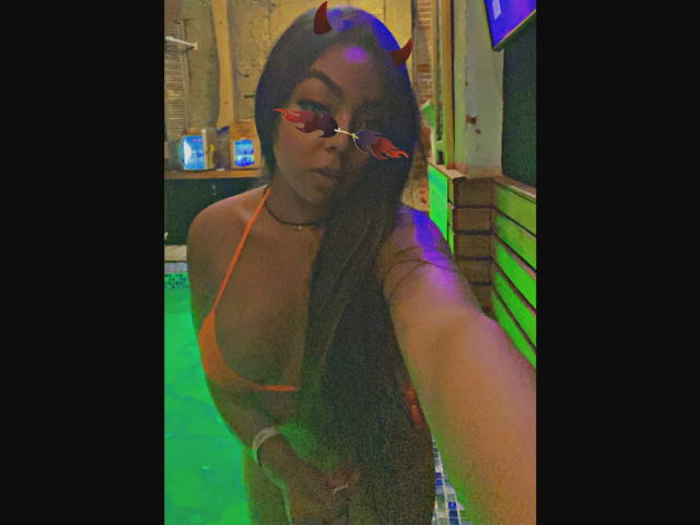 EvaBrown with dark skin, long black hair, orange bikini, red devil horns, and red heart-shaped sunglasses, taking a selfie in a green-lit room. Boobs visible, sexy.