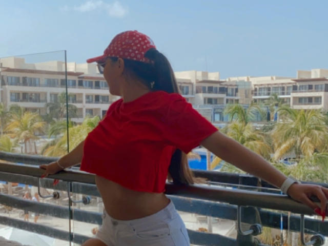EvaBrown, avec une casquette rouge à pois, un crop top rouge, un short blanc et des lunettes de soleil, est appuyée contre une rambarde, montrant son ventre plat et ses petites seins. Sa queue de cheval foncée descend le long de son dos..
