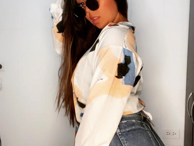 EvaBrown, avec ses longs cheveux bruns, portant des lunettes de soleil foncées, une chemise blanche avec des motifs beige et bleu, et un jean taille haute, montrant ses fesses, regardant par-dessus son épaule..