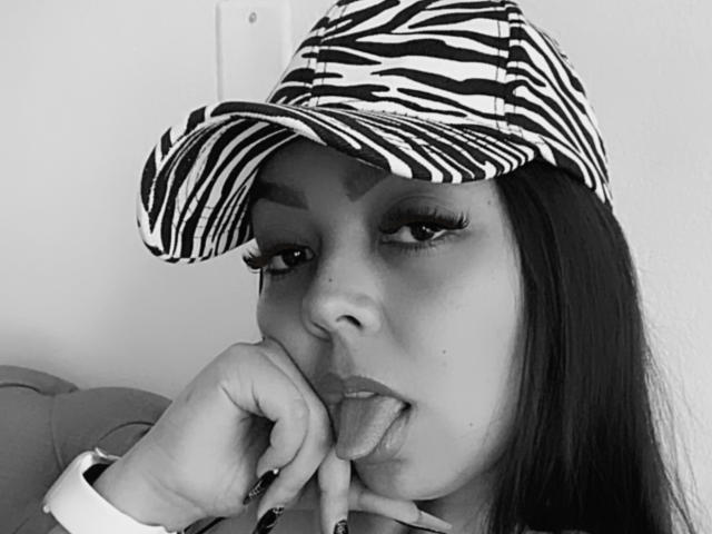 Eva Brown mit einer Mütze mit ZEBRA-Muster, langem, dunklem Haar, dicken Wimpern, die an ihren Fingern lutscht, mit herausgesteckter Zunge, trägt eine Uhr, Schwarz-Weiß-Foto, Fokus auf ihr Gesicht..