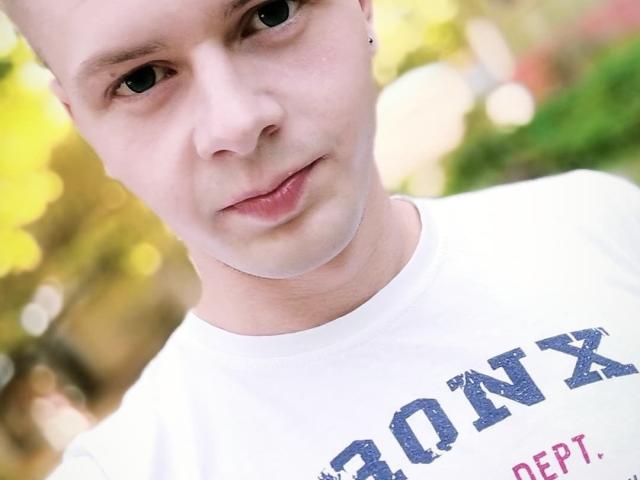 MikeXKing a un visage pâle, un petit nez et un léger sourire. Il porte une chemise blanche avec du texte bleu, et a un petit piercing à l'oreille du côté gauche..