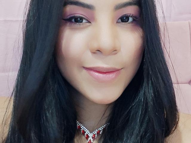 Charlote Evans a les cheveux noirs, un fard à paupières rose et un rouge à lèvres rose, avec un sourire discret, et porte un collier rouge et blanc orné de pierres précieuses, mettant en valeur son visage lisse, mignon et de style asiatique..