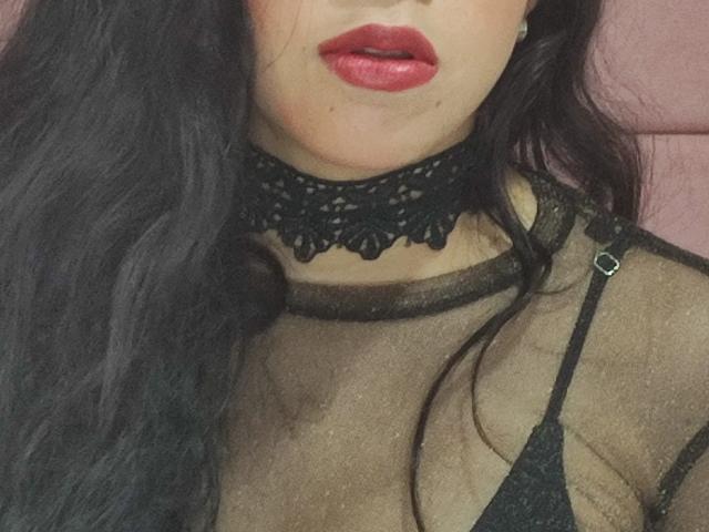 Charlote Evans, com lábios vermelhos, um colar de renda preta e uma blusa preta transparente, mostrando seus seios e uma alça de sutiã preta. Cabelo preto longo e ondulado. Fundo suave e rosado..