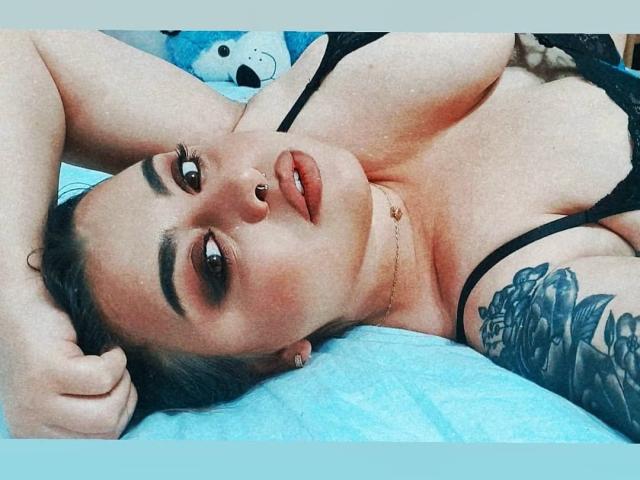 ChaudePourxToi lying on a bed, arms above head, showing tits in a black lace bra, nose ring, tattooed arm, and a sexy look with full lips and dark makeup.