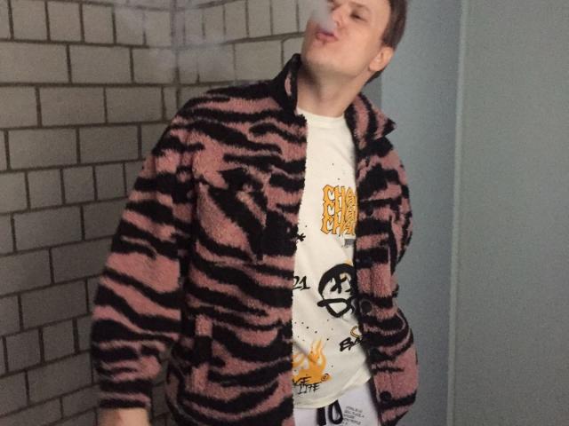 ChillOutt, wearing a pink and black zebra-striped fleece, puffs out a smoke cloud, with eyes half-closed and lips puckered. He's got a white t-shirt with a graphic.