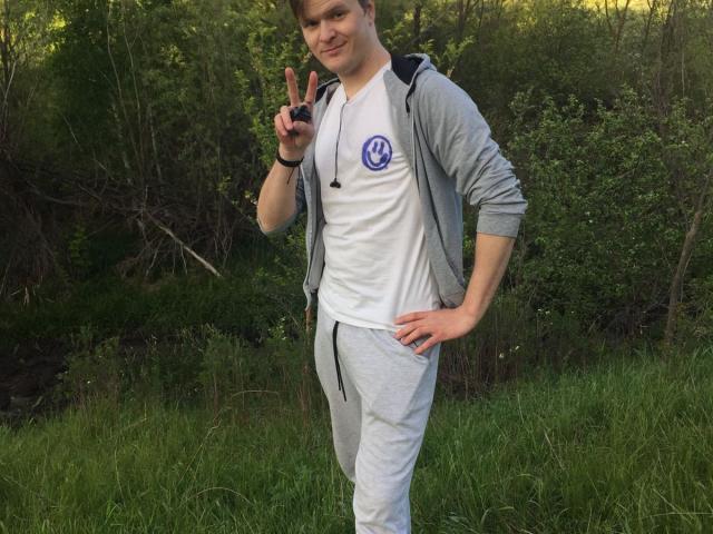 ChillOutt staat in het gras, gekleed in een grijze hoodie, een wit shirt met een VW-logo en grijze joggingbroek. Hij maakt een vrede-teken, met zijn hand op zijn heup, en glimlacht..