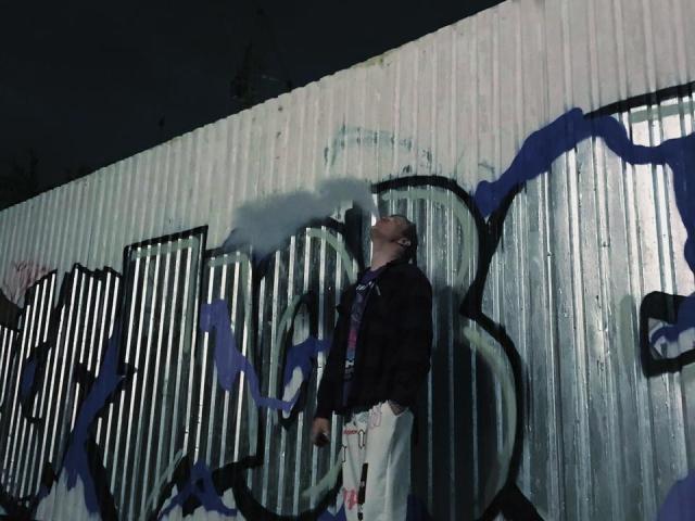 ChillOutt è in piedi di fronte a un graffito, con lo sguardo rivolto verso l'alto e una sigaretta in mano. Indossa una giacca nera, pantaloni bianchi ed è calvo. È notte, l'ambientazione è urbana..