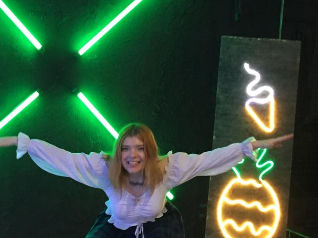 ChillOutt, con cabello rubio, blusa blanca y pantalones negros, con los brazos extendidos, sonriendo, con "X" verdes neón y un letrero de calabaza brillante en el fondo. Tiene una cara linda y feliz..