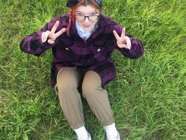 ChillOutt, avec des tresses rousses, des lunettes, un manteau à carreaux violets, un pantalon beige, des chaussettes blanches et des chaussures noires, assise sur l'herbe, faisant deux signes de la paix, souriant..