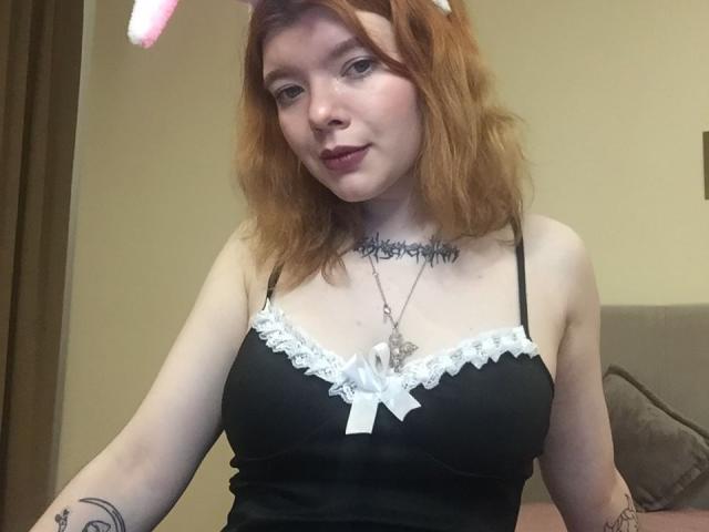 ChillOutt, con i capelli rossi, indossa lingerie nera con pizzo bianco e un fiocco, orecchie da coniglio rosa, rossetto scuro, tatuaggi, una collana e un sorriso carino, mostrando il seno..
