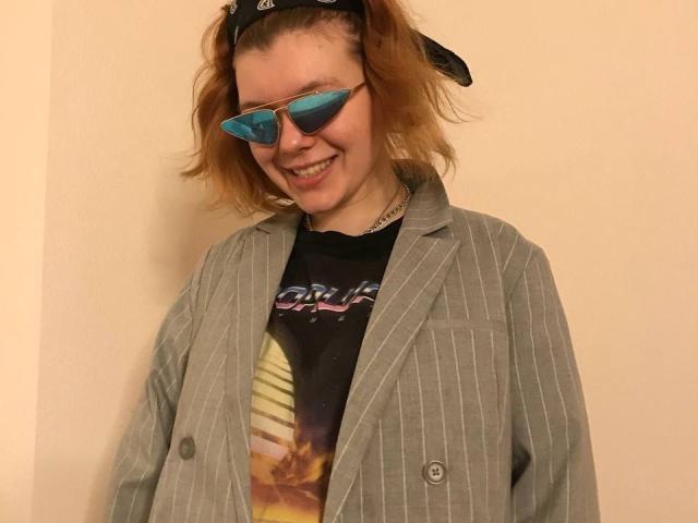ChillOutt, met kort rood haar, blauwe zonnebril met spiegelende glazen, een zwarte bandana, een grijze jas met strepen en een "Star Wars"-shirt, lachend. Ze heeft een tatoeage op haar nek..