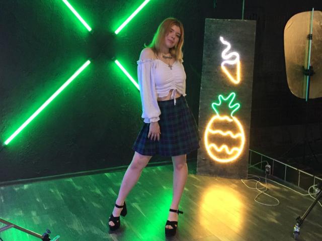 A ChillOutt está em pé, com as pernas afastadas, vestindo uma blusa branca com um ombro descoberto e uma saia verde xadrez, saltos pretos e cabelo loiro. Luzes neon ao fundo..