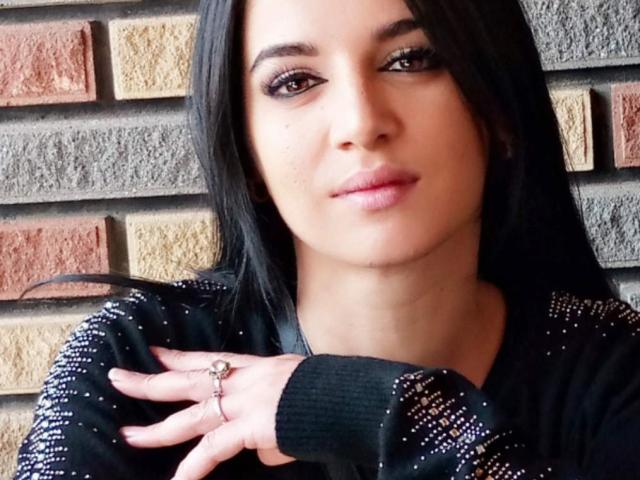 KateLaAngel a de longs cheveux noirs, des yeux foncés et des lèvres roses. Elle porte un pull noir scintillant, avec une bague à sa main droite, et regarde légèrement vers la gauche..