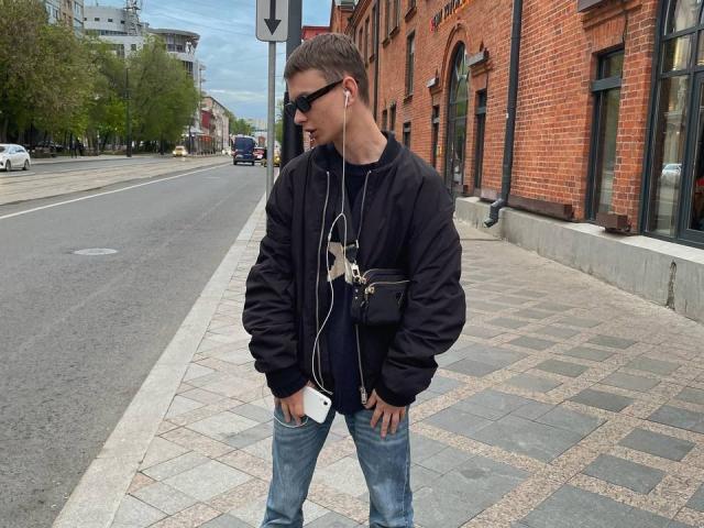 Hombre fumador, con gafas de sol negras, chaqueta negra con calavera blanca, jeans azules y una bandolera negra, de pie en una acera con auriculares puestos, manos en los bolsillos, mirando hacia un lado..