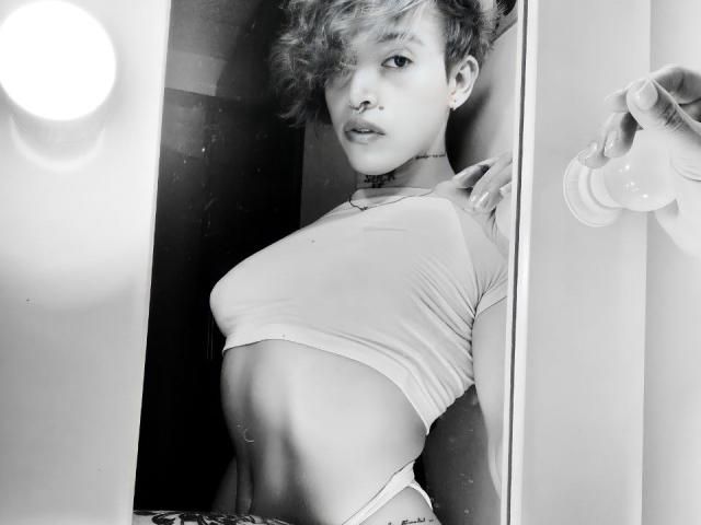 YeiSela with short, messy hair, nose ring, and choker, wearing a tight crop top, showing her small tits and flat stomach, holding a light bulb. Black-and-white photo.