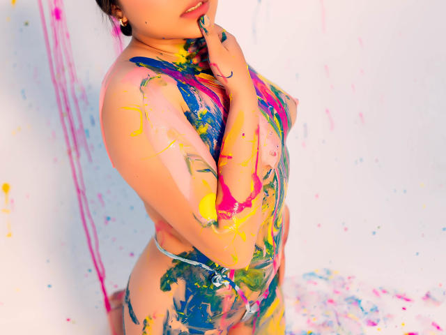 Samantha Velez, nackt, bedeckt mit farbiger Farbe, steht mit der Hand in der Nähe des Mundes, mit bemaltem Busen, farbiger Farbe auf ihrem Gesäß und Oberkörper, weißer Hintergrund, sichtbare Ohrringe, rosa und gelbe Farbe..