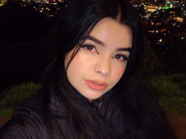 BrithneyLee has pale skin, full pink lips, and dark long hair, looking into the camera with a slight smile. She's wearing a black shirt, with her brown eyes highlighted.