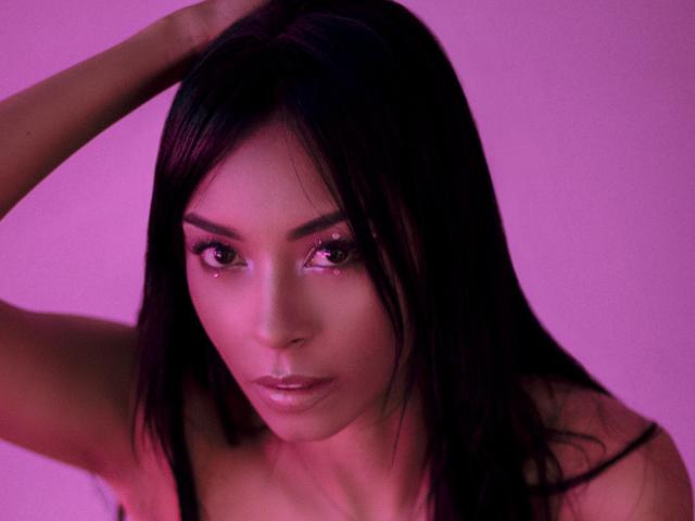 Natasha Holmes, avec ses longs cheveux noirs, son visage illuminé d'une teinte rose et son expression sérieuse, tient ses cheveux en arrière, révélant ses yeux brillants et son maquillage discret. Elle est nue sur le torse, avec ses épaules visibles..