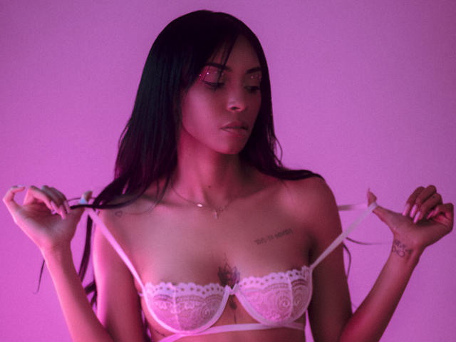 NatashaHolmes with long black hair, pink-lit skin, wearing white lace bra, holding straps, small tattoos on chest, subtle makeup, looking down, pink lighting, soft expression, delicate necklace.