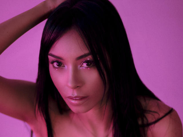 NatashaHolmes has dark hair, purple-lit skin, and piercing eyes looking forward with a serious expression. She's topless, her tits partially visible, with one arm raised.