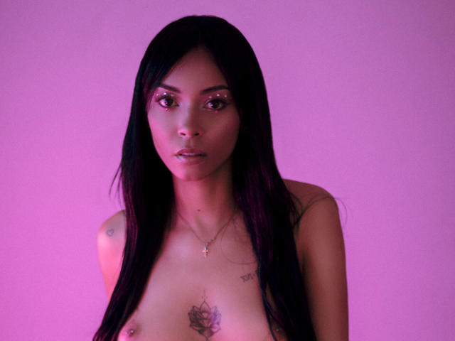 NatashaHolmes with long black hair, pink glittery eyes, topless, showing small tattoos on chest and upper arms, pink glow, looking at camera, subtle necklace, serious expression.