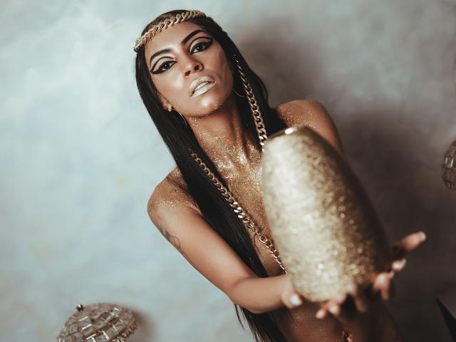 Natasha Holmes, avec une peau brillante et scintillante, tenant un objet doré et phallique dans sa main, portant une coiffe en or et de longues tresses noires, regardant séductrice l'appareil photo..