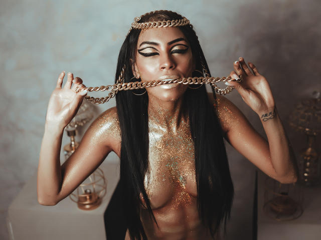 NatashaHolmes with long black hair, golden chain in mouth, glittering skin, nude, holding chain with hands, wearing headpiece, eyes closed, shiny gold makeup, visible small breasts, sexy.