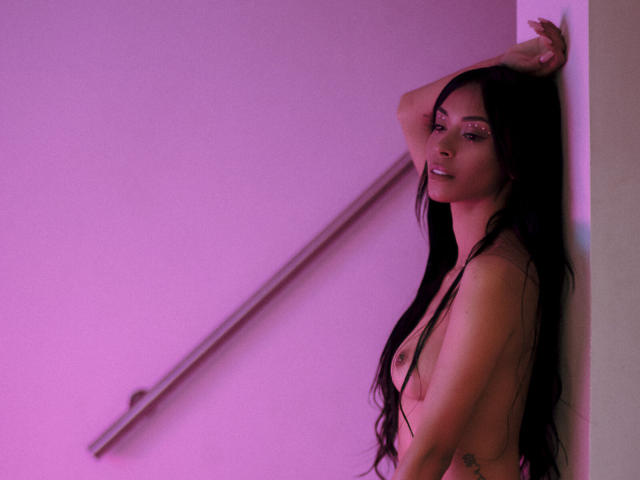 NatashaHolmes with long black hair, topless, standing against a wall, right arm raised, pink-tinged lighting, side profile, visible small tits, subtle tattoo on right side.