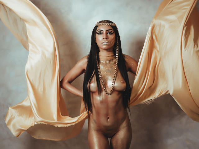 NatashaHolmes with long black hair, wearing gold jewelry, and a headpiece, standing naked with flowing golden fabric, exposing her tits and shiny gold-covered skin, arms behind her back.