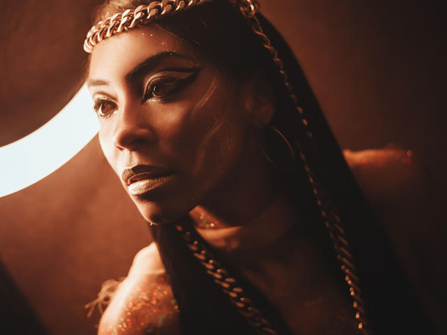 NatashaHolmes with dark skin, braids, gold chain headpiece, bold eyeliner, and shiny makeup, looking serious and sexy, illuminated by warm light. Boobless, but very attractive.