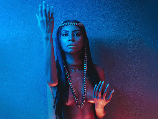 NatashaHolmes with long braids, wearing a chain headpiece and chain necklace, topless, pressing hands against a blue and red lit wall, looking serious, with a silver chain on her tits.
