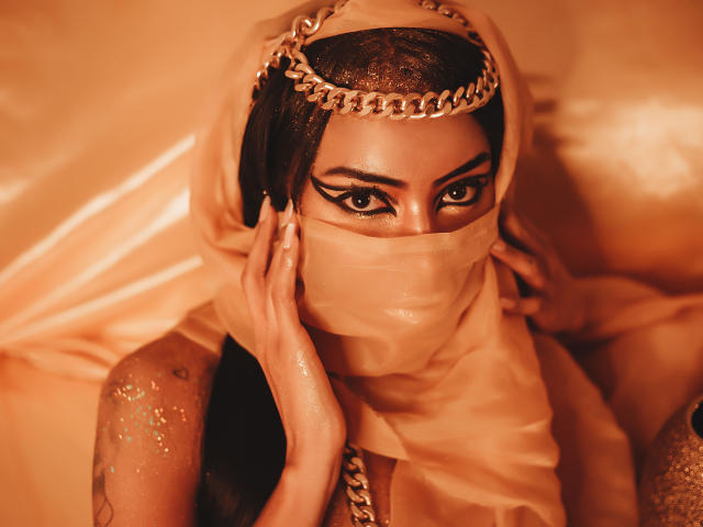 Natasha Holmes, à la peau foncée, portant une coiffe en or, un masque beige et un tissu drapé, a des bras brillants, mouillés et scintillants, et des yeux intenses et séduisants, levés vers le haut..