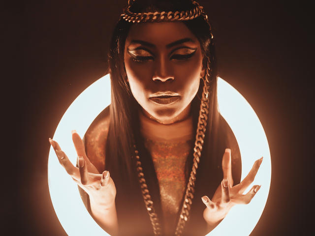 NatashaHolmes with long dark hair, gold chain headpiece, and glowing circular light behind. She's making a "rock on" gesture with both hands, looking down with a serious expression.