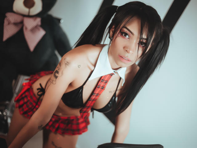 Natasha Holmes, avec ses couettes noires, portant une jupe à carreaux rouges, un soutien-gorge noir et une chemise blanche, a des tatouages et est penchée en avant, montrant ses seins et ses fesses..