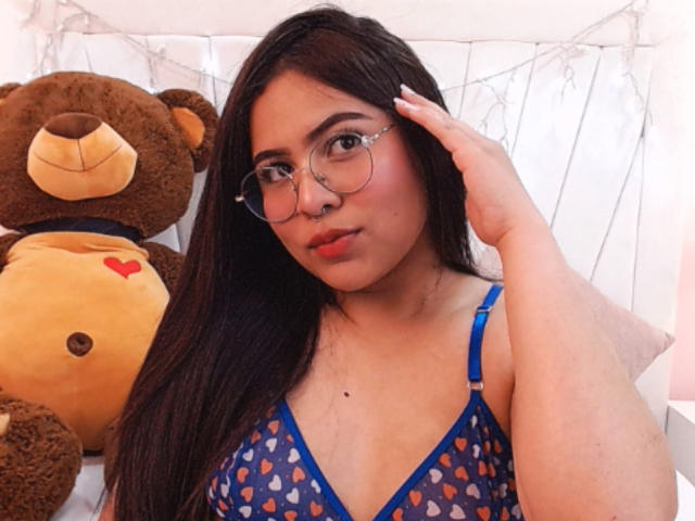 Samantha Velez mit langen, dunklen Haaren, Brille, Nasenring, einem blauen BH mit Herzmuster, einer Hand auf dem Kopf, einem braunen Teddybären mit Herz auf der Brust, bei weicher Beleuchtung..