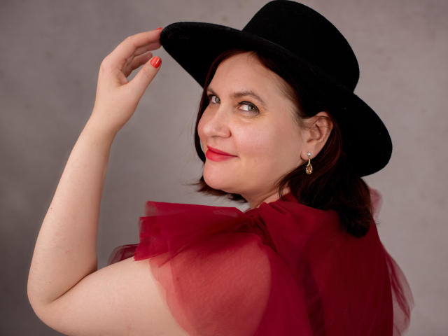 BigTitsXHot with red tulle dress, black hat, and earrings, raising her hand with red nails, smiling at the camera, showing a cute face and short brown hair.