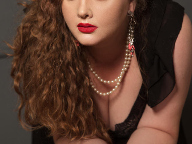 SinFulTitsDD con cabello castaño rizado, lápiz labial rojo, collar de perlas, blusa de encaje negro y aretes rojos, inclinándose hacia adelante, mostrando sus pechos..