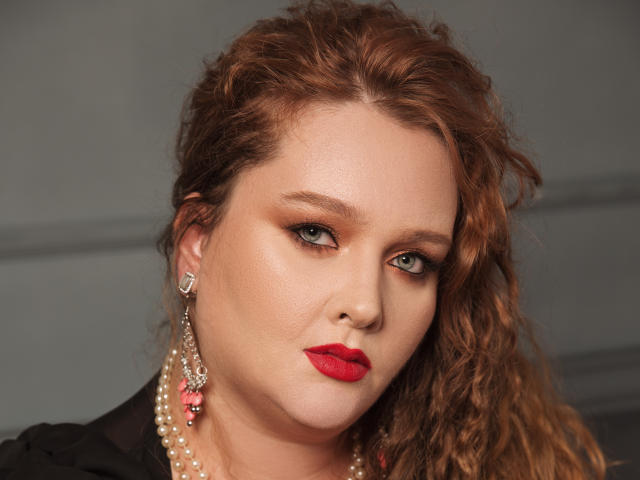 SinFulTitsDD has curly red hair, red lipstick, and pearl earrings with pink gems. She's looking slightly to the side with a serious expression. She has fair skin and green eyes.