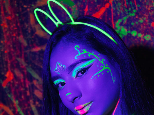 MiaSuarez with glowing neon bunny ears, blue face paint, and neon green makeup, smiling under colorful lights, her long dark hair illuminated, looking cute and playful.