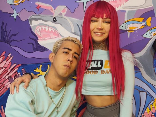 HardCoreSexy, com cabelo ruivo vibrante, tatuagens e um piercing no nariz, está ao lado de um menino loiro que veste uma camisa azul clara. Ela está usando uma cropped top, enquanto ele veste um moletom..