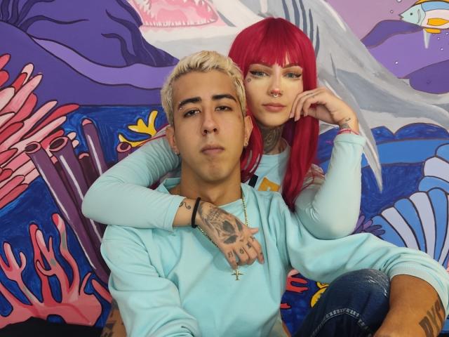 HardCoreSexy mit blonden Haaren und Tätowierungen, die ein hellblaues Hemd trägt, sitzt vor HardCoreSexy mit roten Haaren, die ebenfalls ein hellblaues Hemd trägt und sich an ihn lehnt..