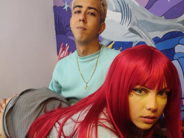 HardCoreSexy with red hair, eyeliner, nose ring, and gold necklace leans forward, looking at camera, while HardCoreSexy with blonde hair, blue shirt, and gold necklace sits behind.