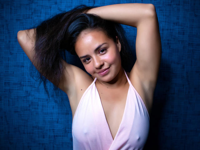 ArielleWilde with arms up, dark hair, smiling, pink halter top, showing small tits, armpits, and a cute face, against a blue textured background.