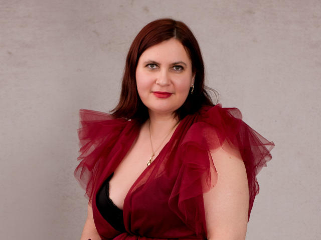 BigTitsXHot with red hair, wearing a deep red, ruffled dress with black bra showing, smiling at camera, gray background, subtle makeup, gold necklace, looking cute and sexy.