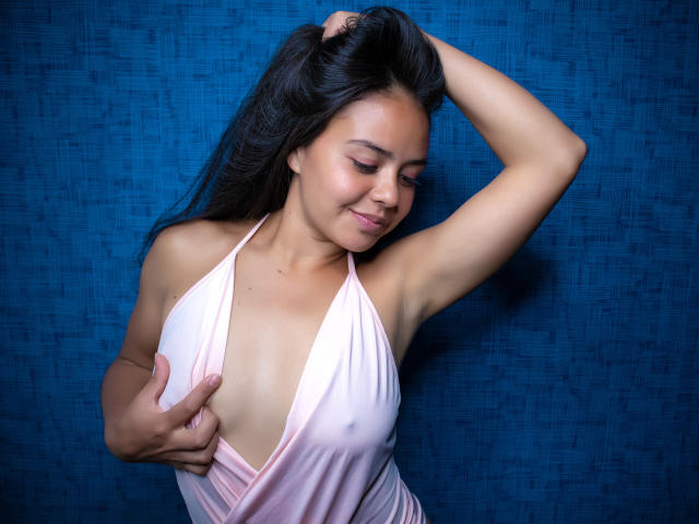 ArielleWilde with tan skin, dark hair, and small tits, wearing a pink halter dress, holds her hair up, eyes closed, smiling, hand on her chest, blue background.